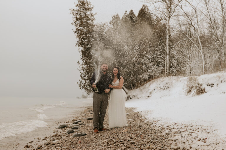 Eloping couple popping champagne to celebrate on snowy lakefront