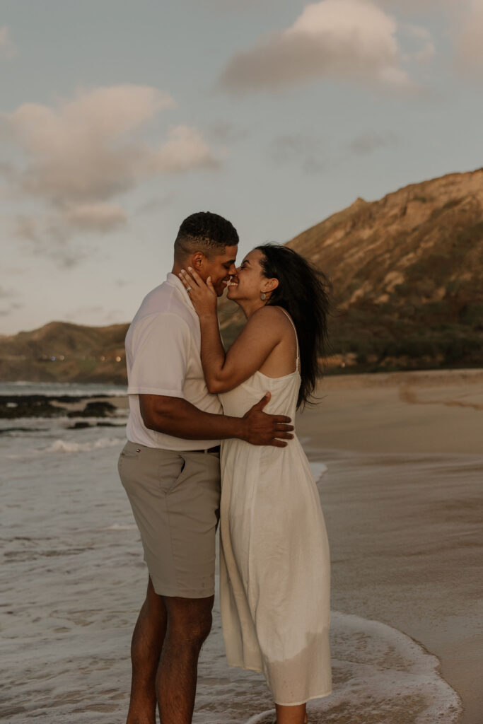 couple kissing on the beach at sunrise in Oahu, Hawaii with mountains in the background