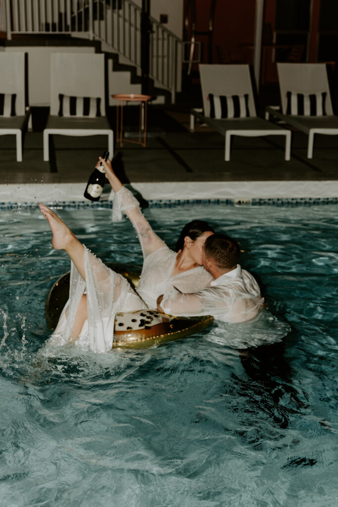 Couple jumping into a pool with champagne in their wedding clothes