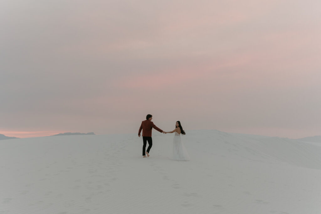 Couple running in white sand dunes toward pink skies